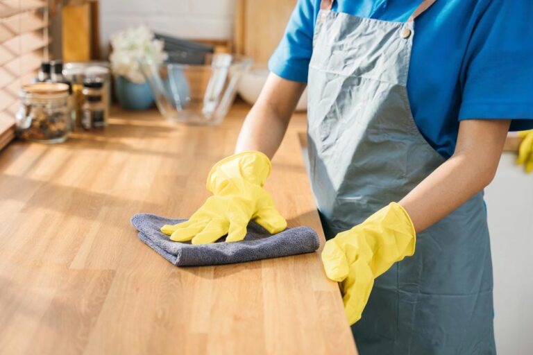 Maid in protective glove cleans wood table in cozy kitchen. Using professional cleaning products for home perfect tidiness. Cleaner working safety glove hygiene routine. Maid housekeeping concept.
